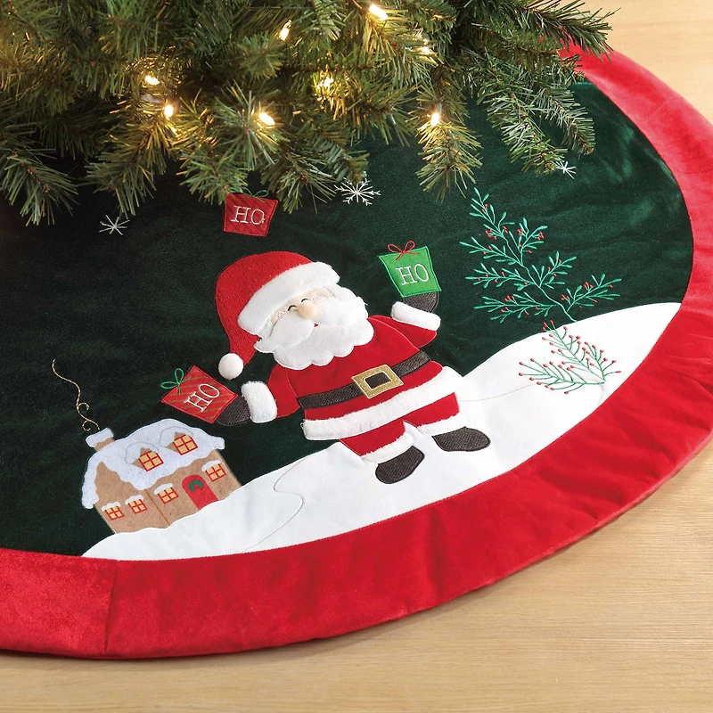 Jupe d'arbre de Père Noël en velours, 48 pouces, multi couleur Jupe d'arbre de Noël,matériau de velours,couleur vert foncé de base,couleur rouge de garniture,le Père Noël se tient sur le sol enneigé,tenant des sacs cadeaux verts et rouges avec "HO" brodé sur lui.Il y a des fleurs sur le sol enneigé,une maison au dos du Père Noël.