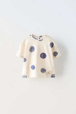 Round neck T-shirt with short sleeves. Label appliqué at hem. Buttoned teardrop closure back.