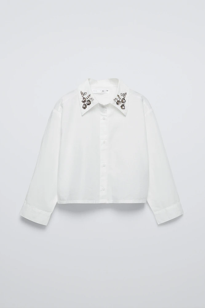 Shirt with collar, long sleeves, and buttoned cuffs. Front button closure. Jewels appliqué at collar.