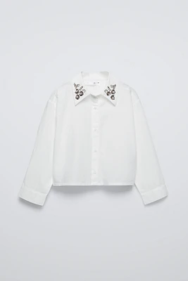 Shirt with collar, long sleeves, and buttoned cuffs. Front button closure. Jewels appliqué at collar.