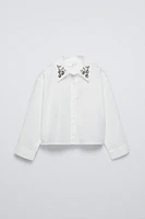 Shirt with collar, long sleeves, and buttoned cuffs. Front button closure. Jewels appliqué at collar.