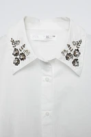 Shirt with collar, long sleeves, and buttoned cuffs. Front button closure. Jewels appliqué at collar.