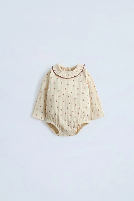 Round neck bodysuit with ruffle appliqué and long sleeves. Snap button closure at back bottom. Cherry print.