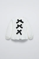 Shirt with round neck and long sleeves. Buttoned teardrop closure at back. Contrasting front bows appliqué. Ruffled hem.