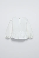 Shirt with round neck and long sleeves. Buttoned teardrop closure at back. Contrasting front bows appliqué. Ruffled hem.