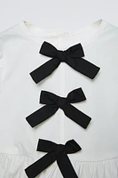 Shirt with round neck and long sleeves. Buttoned teardrop closure at back. Contrasting front bows appliqué. Ruffled hem.